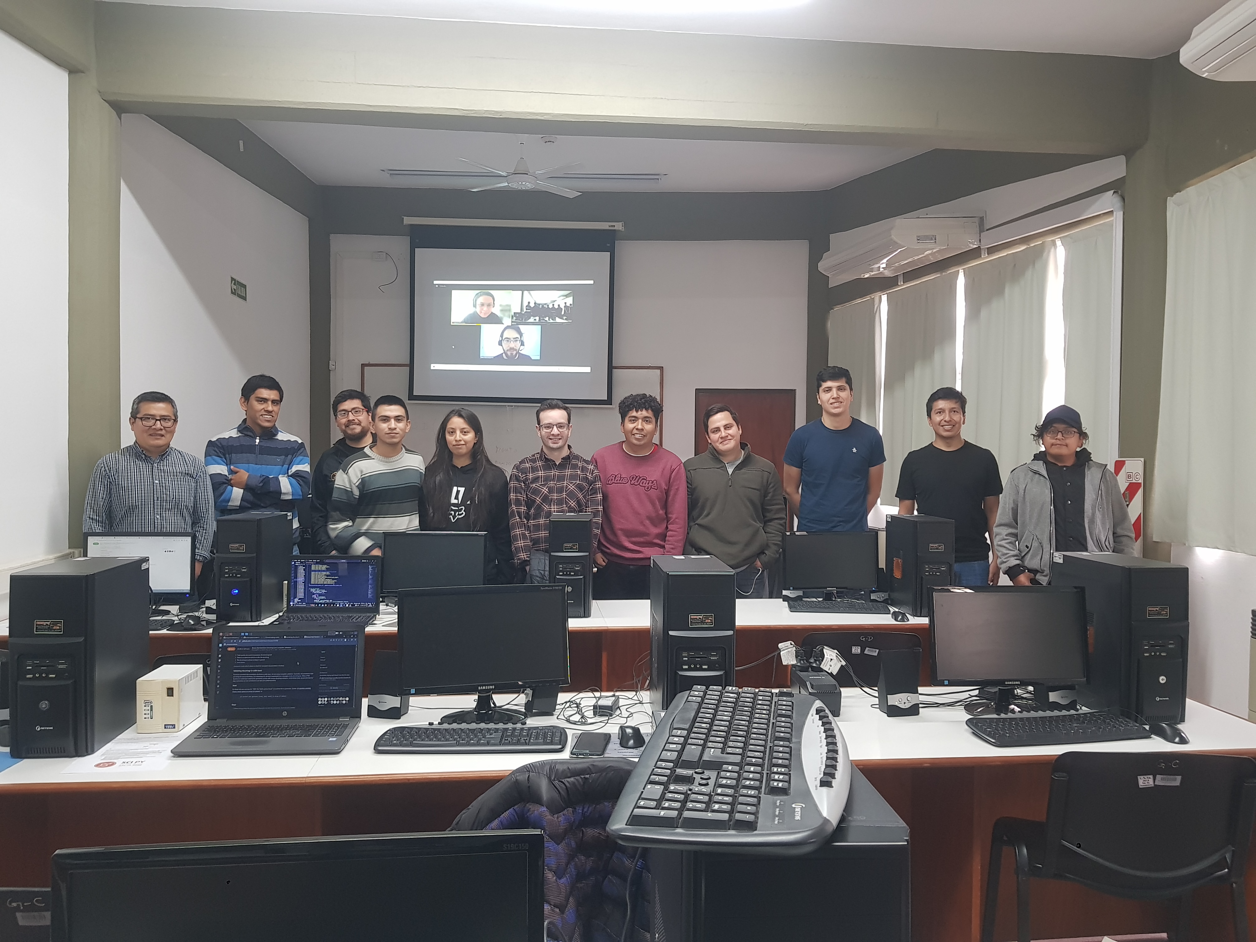 11 people standing behind some computers and 2 people projected in the screen